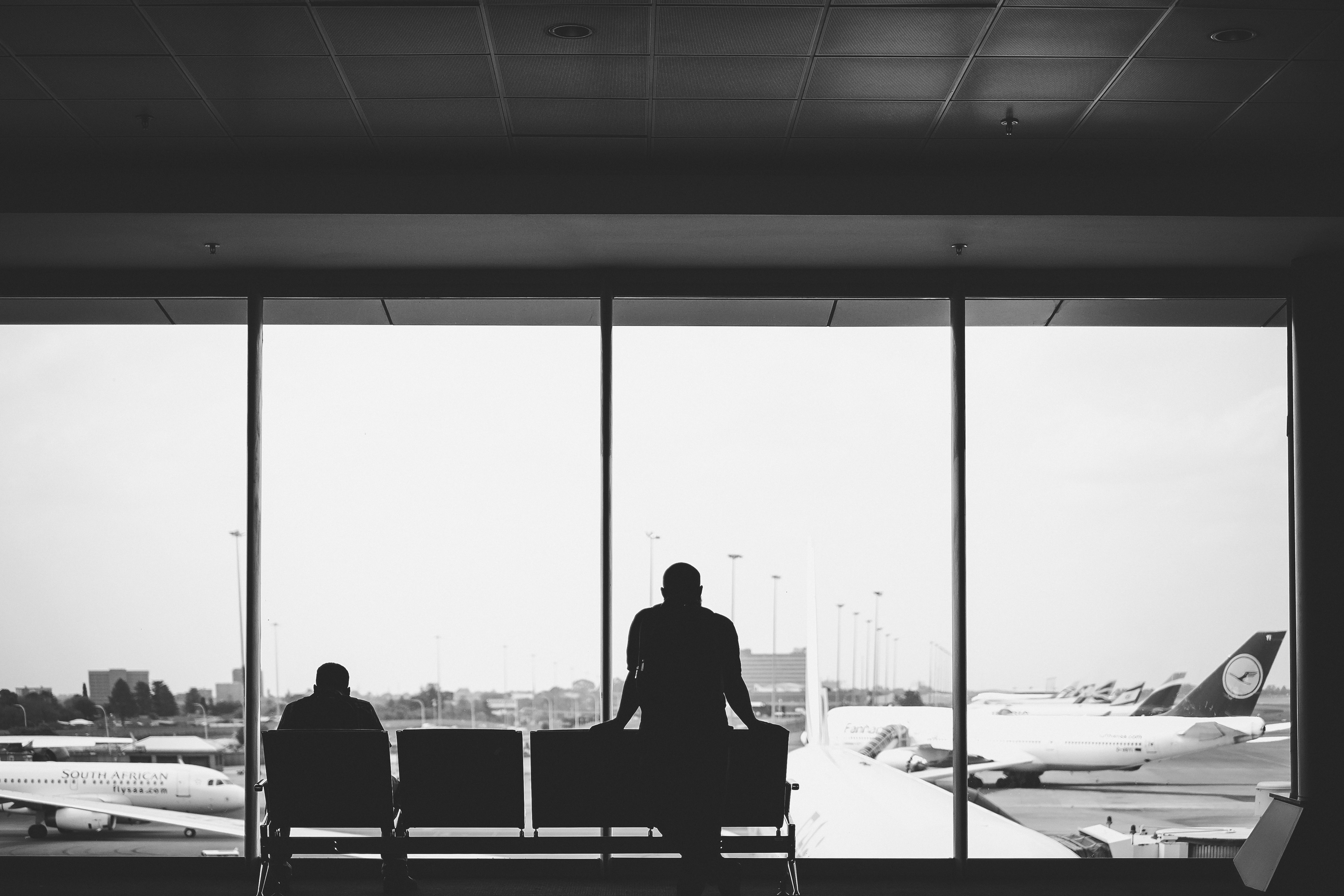 To personer venter inne på en flyplass. Utenfor vinduet er det flere fly som står på rekke og rad. Foto.